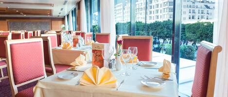 A restaurant with tables set for dining, featuring a view of buildings and trees through large windows.