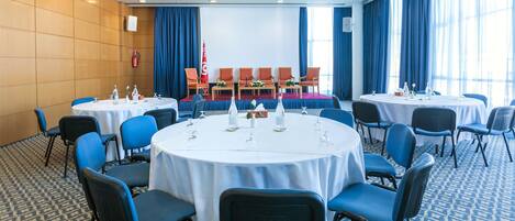 A conference room with round tables, blue chairs, and a stage with a podium and chairs.