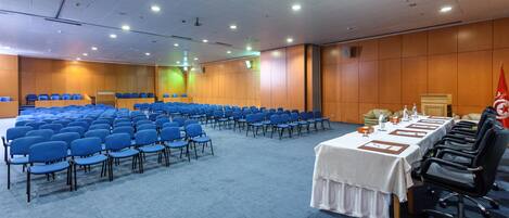 Une salle de conférence avec des rangées de chaises bleues, une longue table ornée de plaques nominatives et un pupitre.