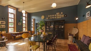 Una habitación con una gran mesa de madera, varias sillas, un sofá y un banco. Hay ventanas con cortinas, una planta en maceta y obras de arte colgadas en la pared.