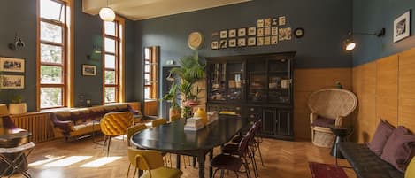 A room with a large wooden table, multiple chairs, a sofa, and a bench. There are windows with curtains, a potted plant, and wall-mounted artwork.