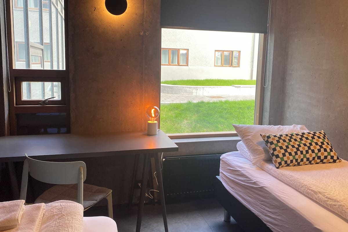 A bedroom with a bed, a desk, and a chair. There is a window with a view of a building and a lawn.