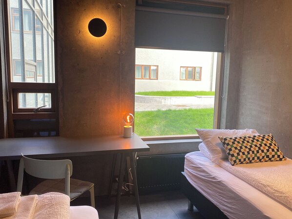 Un dormitorio con cama, escritorio y silla. Se ve una ventana con vistas a un edificio y un jardín.