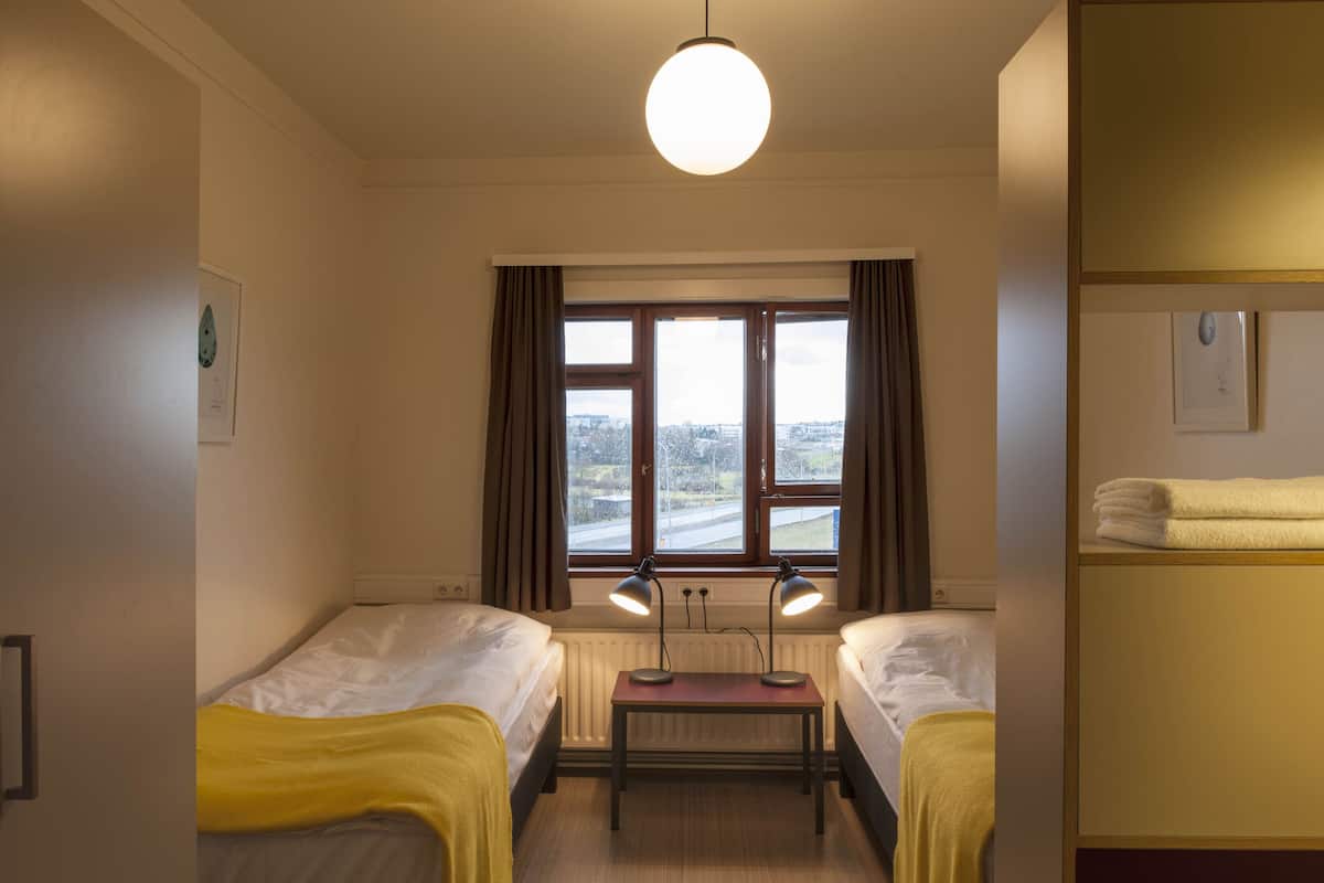 A room with two single beds, a small table, a window with curtains, and a radiator.