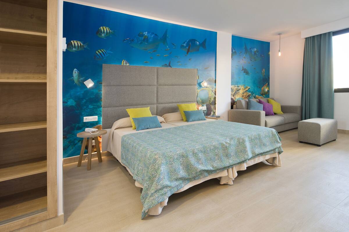 A hotel room with a bed, a sofa, a small table, and a wall mural of fish.