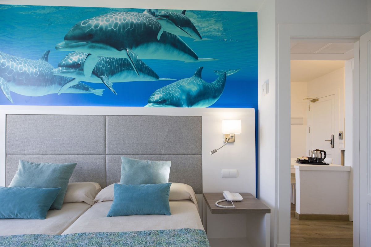 A hotel room with a bed, two pillows, a bedside table with a phone, and a wall mural of dolphins.
