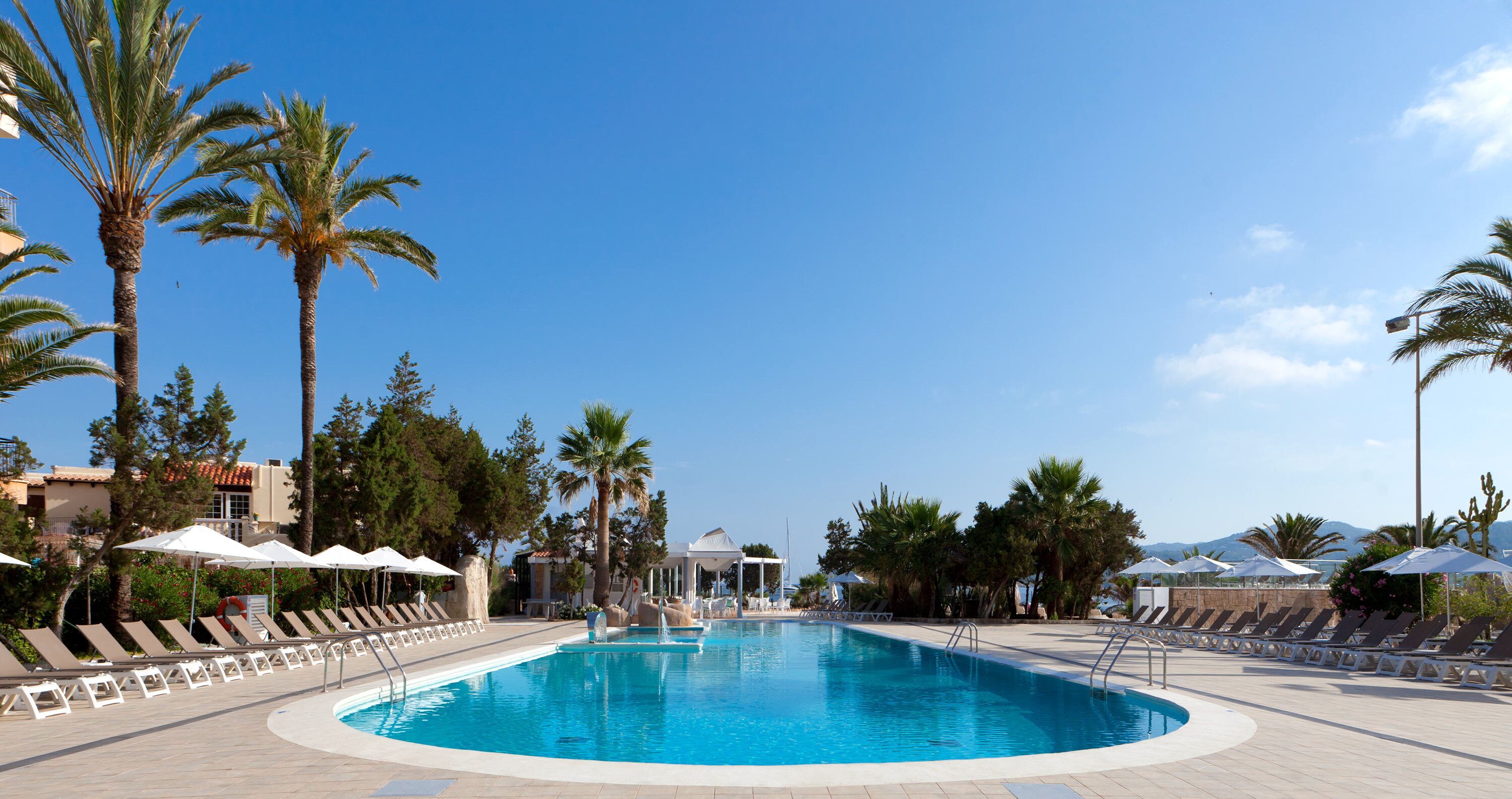 Zona de piscina con sillones de descanso y sombrillas, rodeada de palmeras y un cielo despejado.