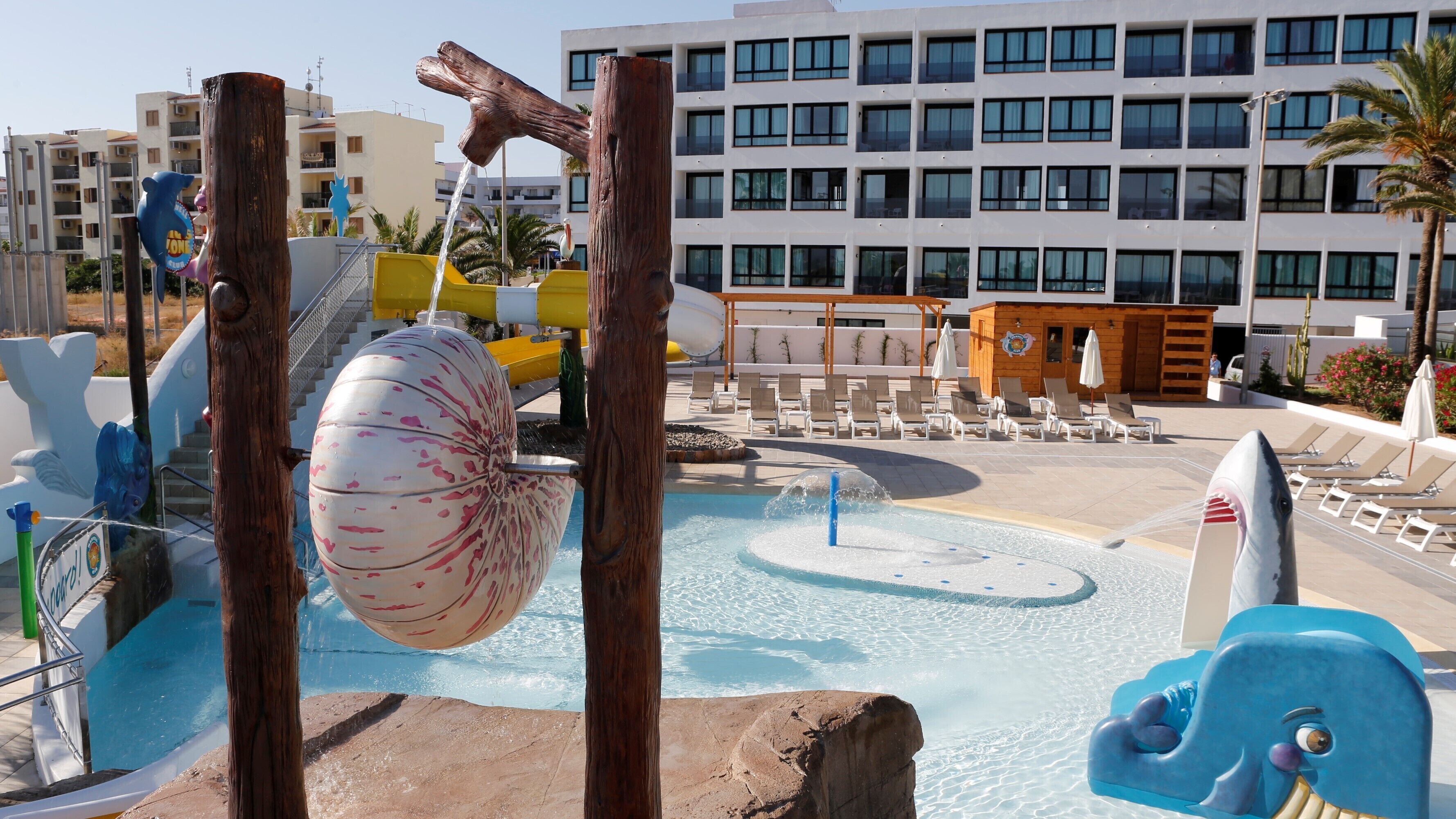 Zona de piscina con elementos acuáticos, incluyendo una noria grande y una estructura de juegos con forma de tiburón. Hay sillones de descanso y un edificio con múltiples ventanas al fondo.