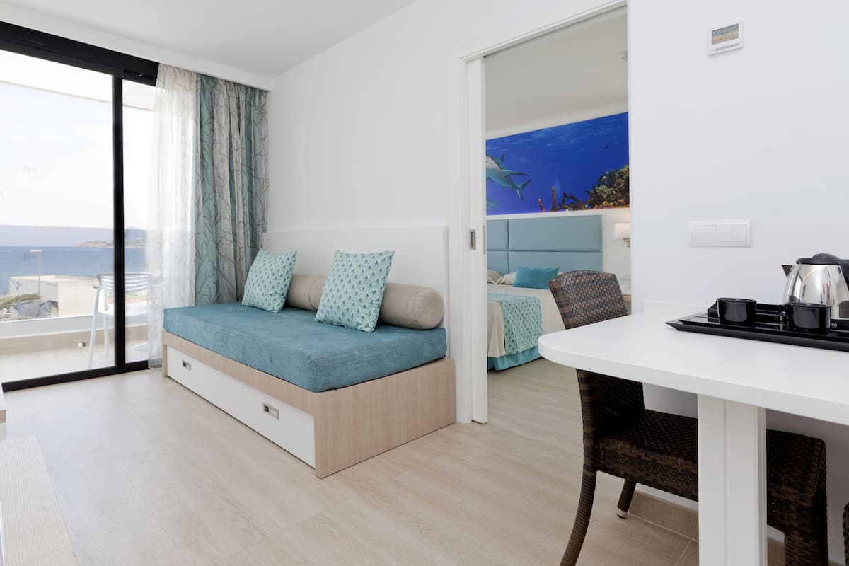 A modern hotel room with a bed, a dining area, and a view of the sea.