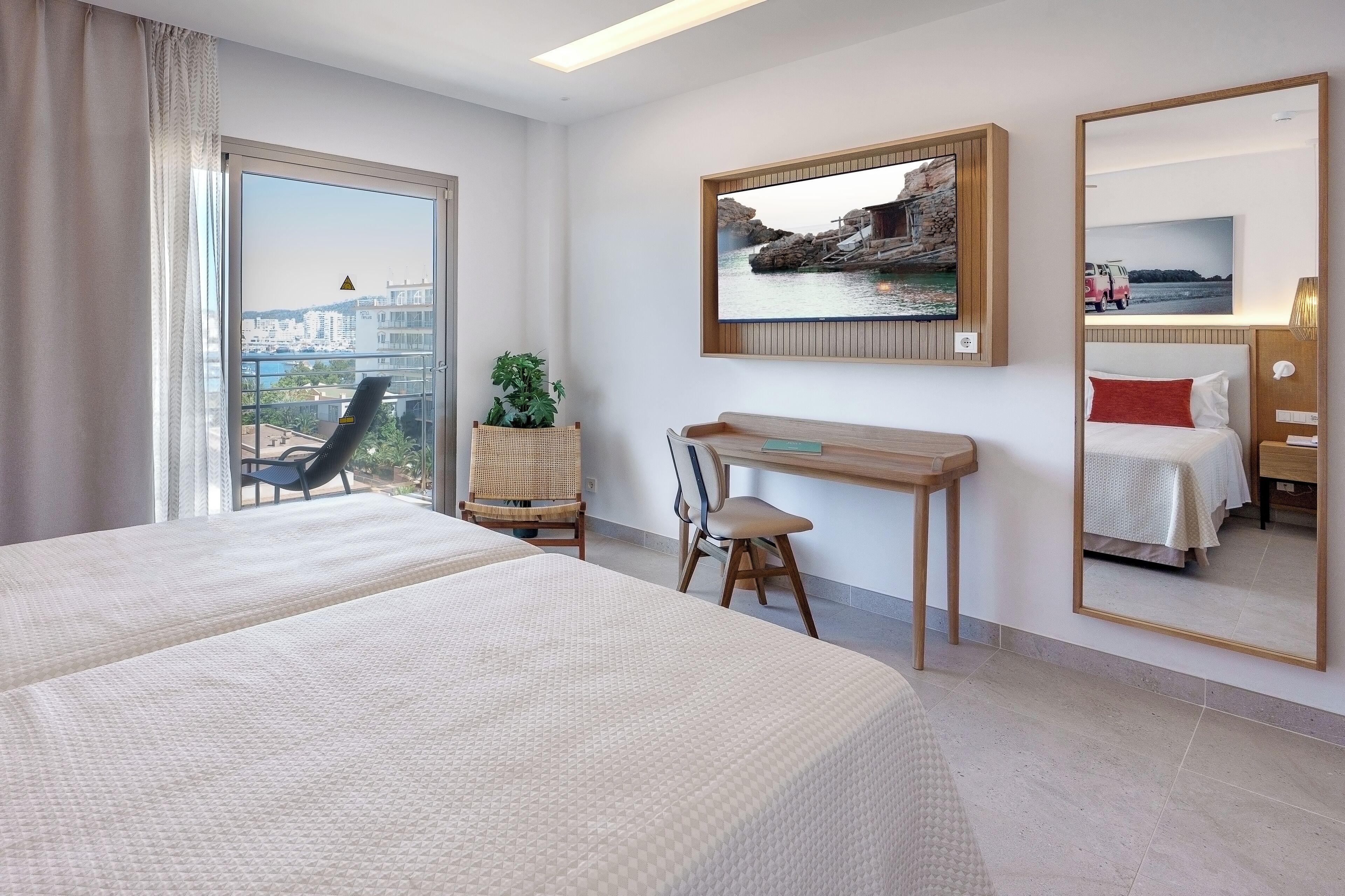 A hotel room with a large bed, a desk, a chair, a potted plant, a large mirror, and a view of the city through a window.