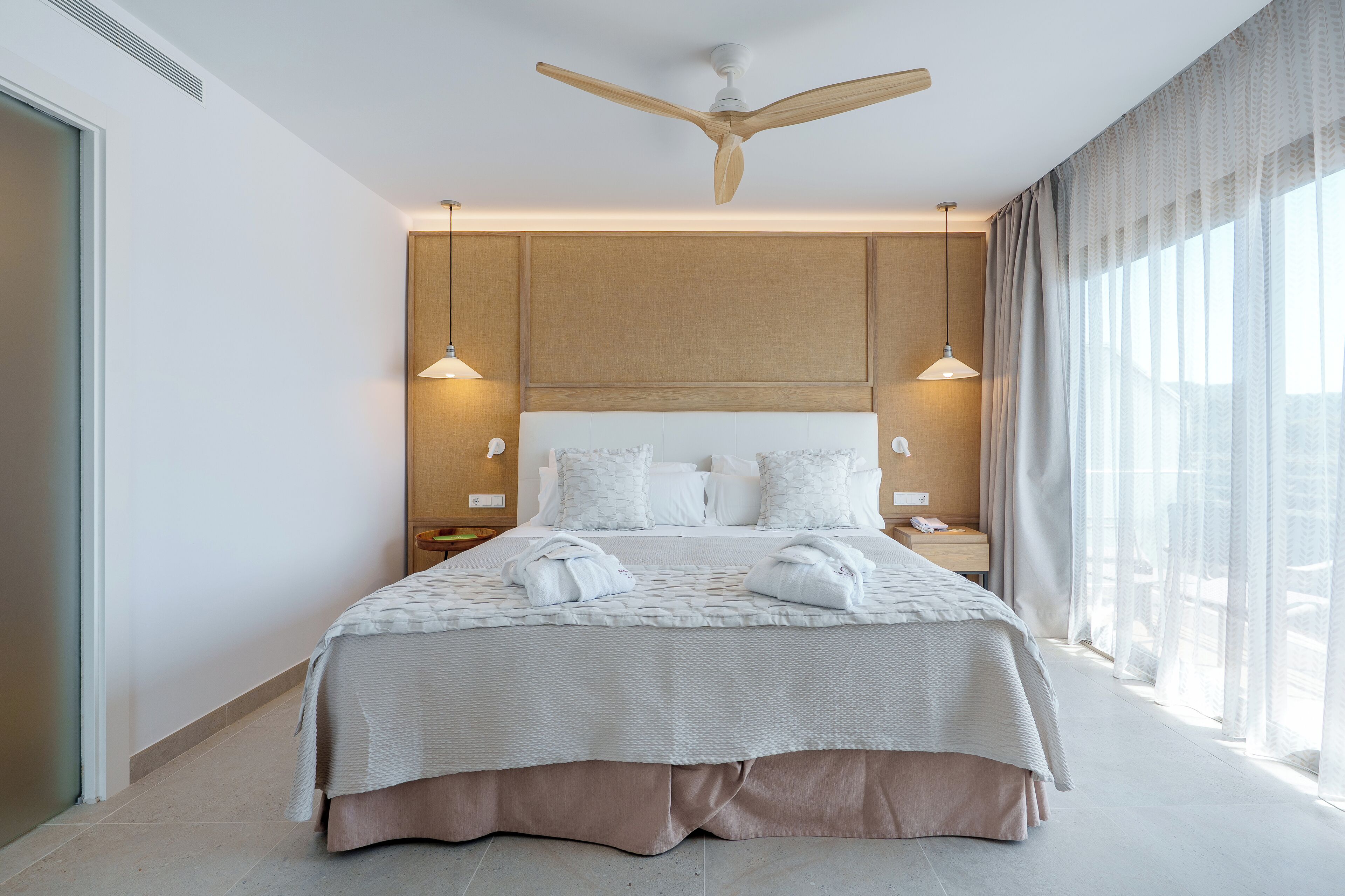 A modern hotel room with a large bed, a ceiling fan, and a view of the city through sheer curtains.