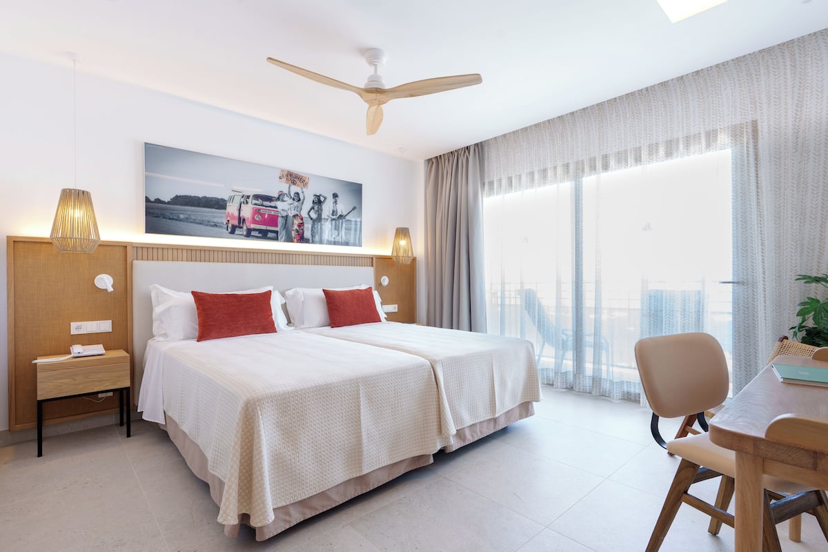 A hotel room with a large bed, a desk, and a chair. There is a ceiling fan, a painting on the wall, and a window with curtains.