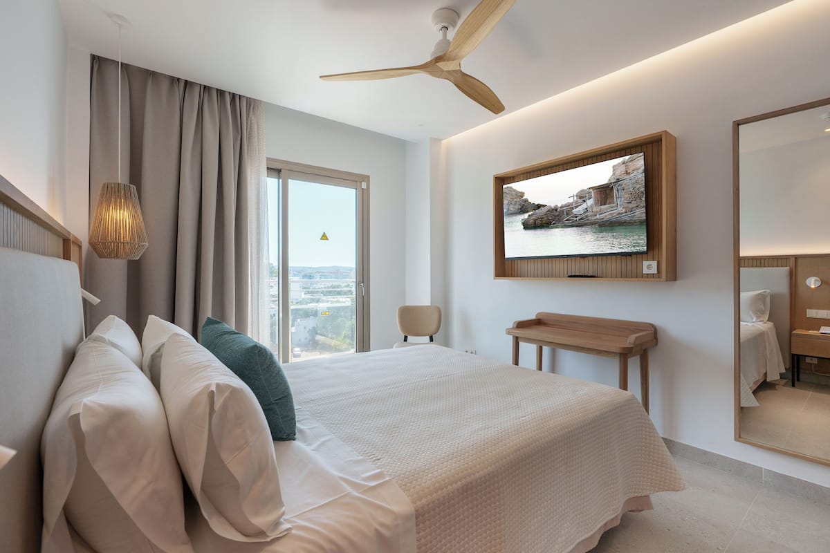 A modern hotel room with a large bed, a ceiling fan, a television, and a view of the city through the window.