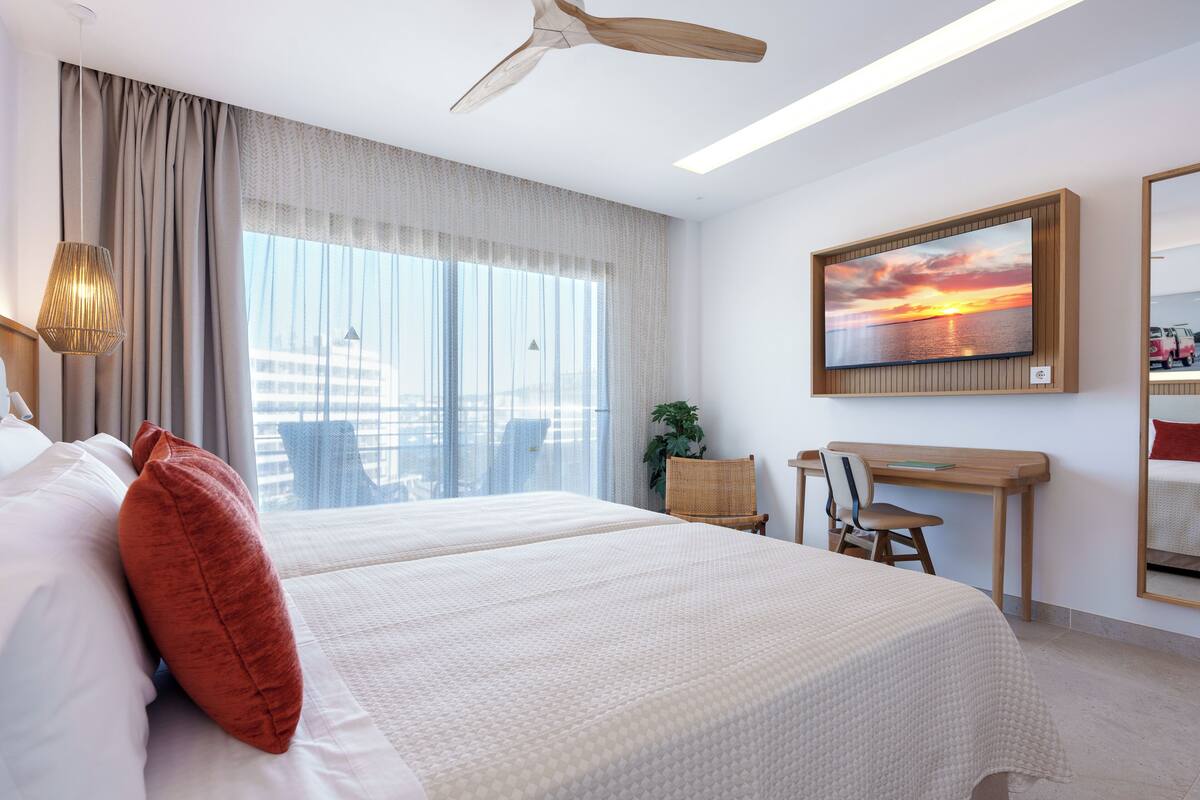 A hotel room with two beds, a desk, a chair, a large window with curtains, a ceiling fan, and a painting of a sunset.
