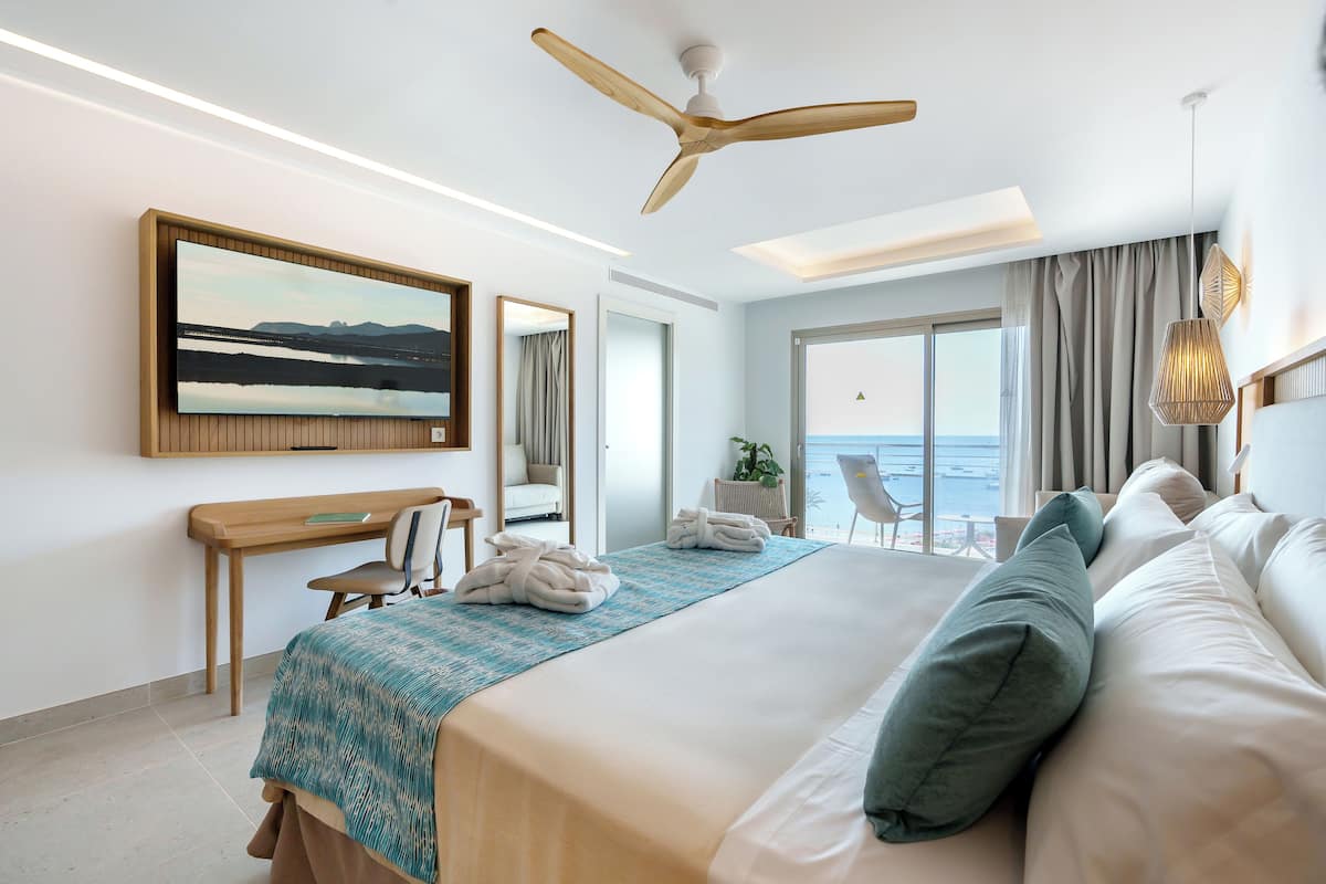 A modern hotel room with a large bed, a desk, a chair, a ceiling fan, and a view of the sea through the window.