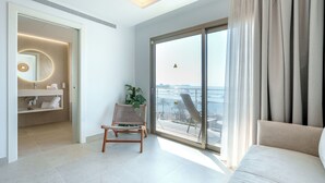 Een moderne hotelkamer met een grote schuifglazen deur naar het balkon, een zithoek met een bank en een fauteuil, en een badkamer met een ronde spiegel en een handdoekhouder.