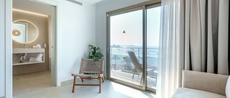 Una habitación de hotel moderna con un amplio ventanal corredero al balcón, una zona de estar con sofá y sillón, y un baño con espejo circular y toallero.