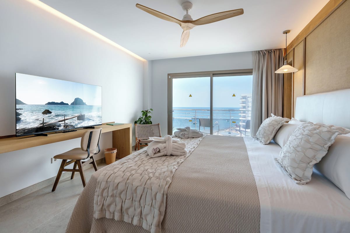 A modern hotel room with a large bed, a TV on the wall, a desk with a chair, and a view of the sea through the window.