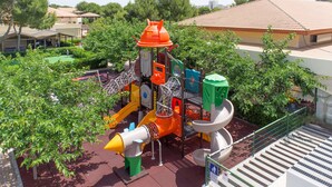 A playground with various play structures, surrounded by greenery and a building in the background.