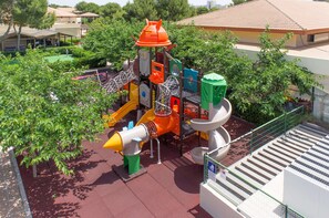 A playground with various play structures, surrounded by greenery and a building in the background. - Zafiro Mallorca (Santa Margalida)