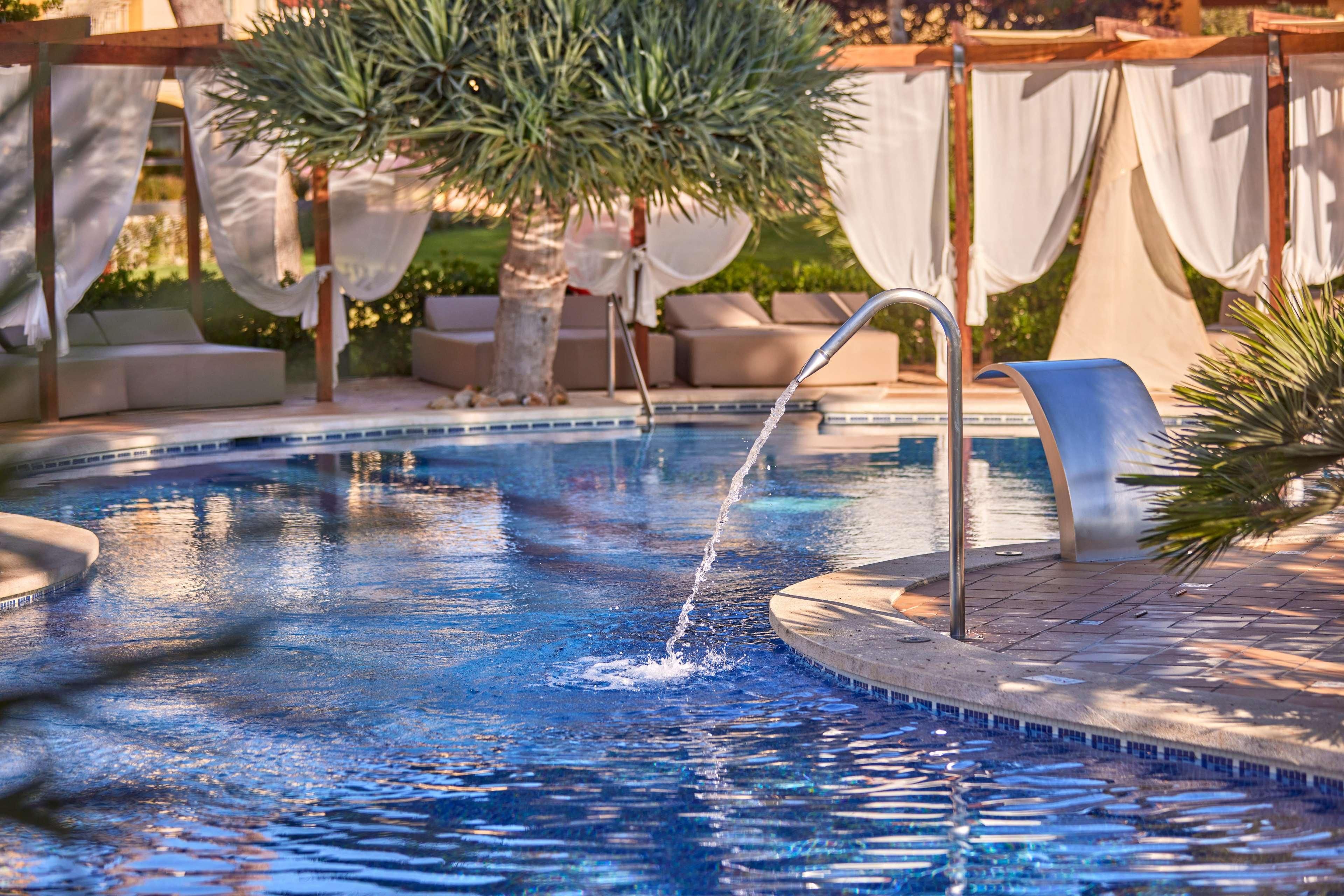 A swimming pool with a water feature, surrounded by a pergola with white curtains.
