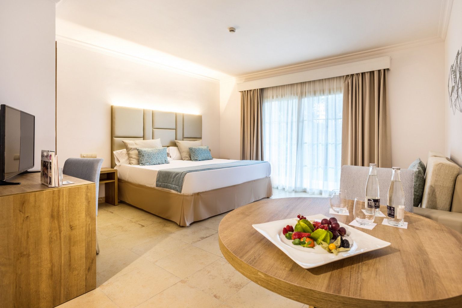 Habitación de hotel con cama, escritorio, mesa redonda con fruta y vasos, y televisor a la izquierda.