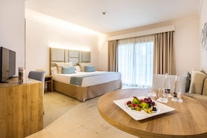 Habitación de hotel con cama, escritorio, mesa redonda con fruta y vasos, y televisor a la izquierda.