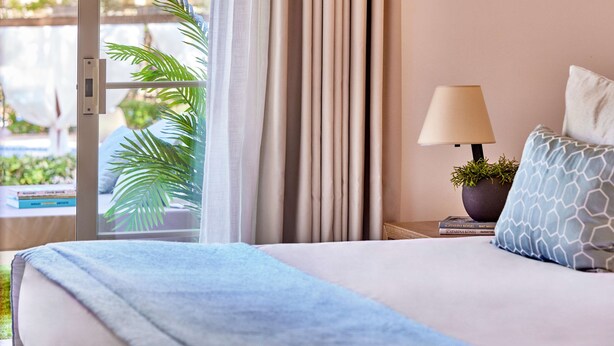 A hotel room with a bed, a nightstand, a lamp, a plant, and a view of a pool.