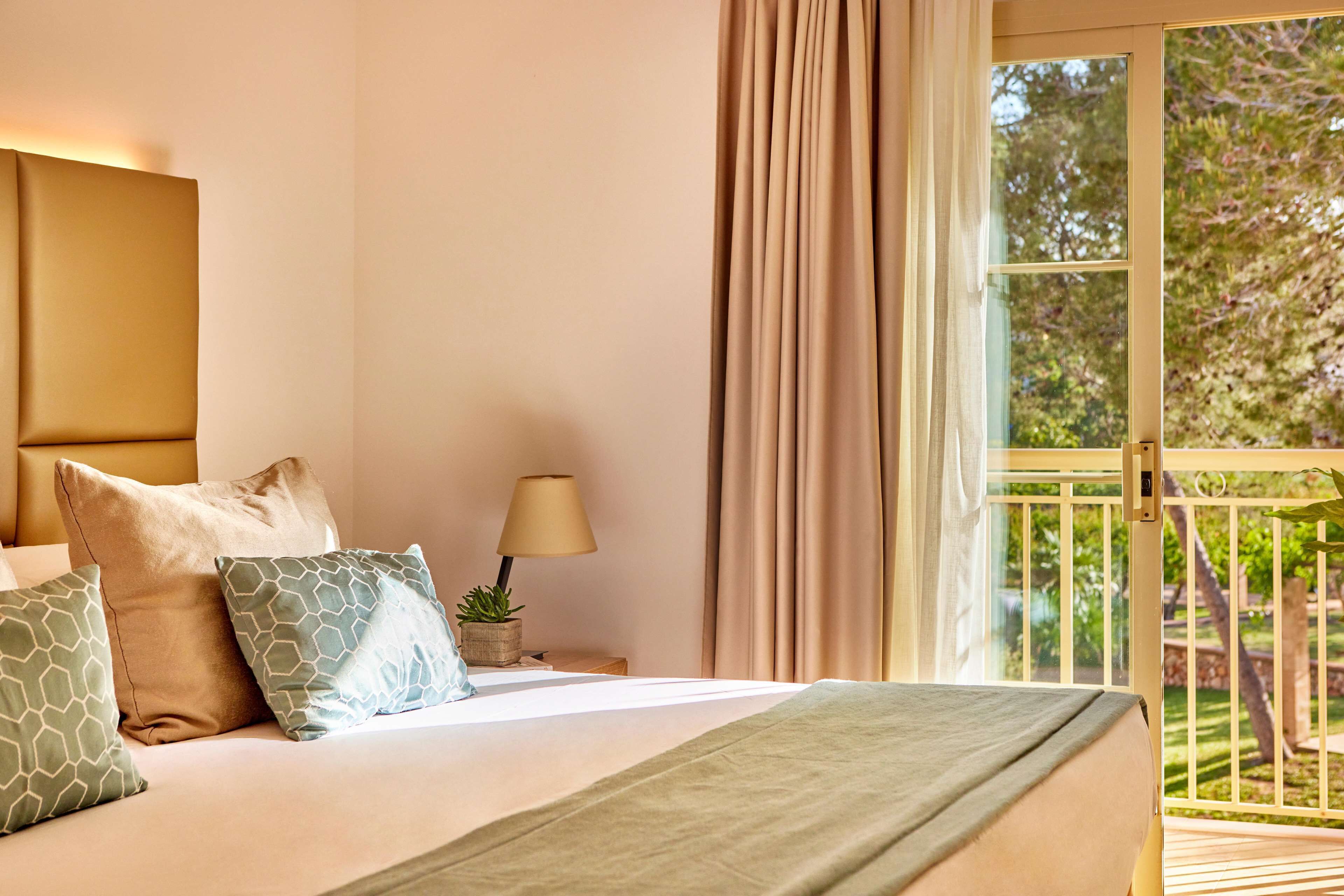 A bedroom with a bed, pillows, a nightstand with a lamp, and a view of a balcony.
