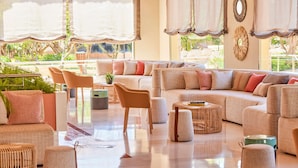 A spacious living area with a beige sofa, multiple chairs, and a coffee table. There are potted plants and a view of greenery outside.