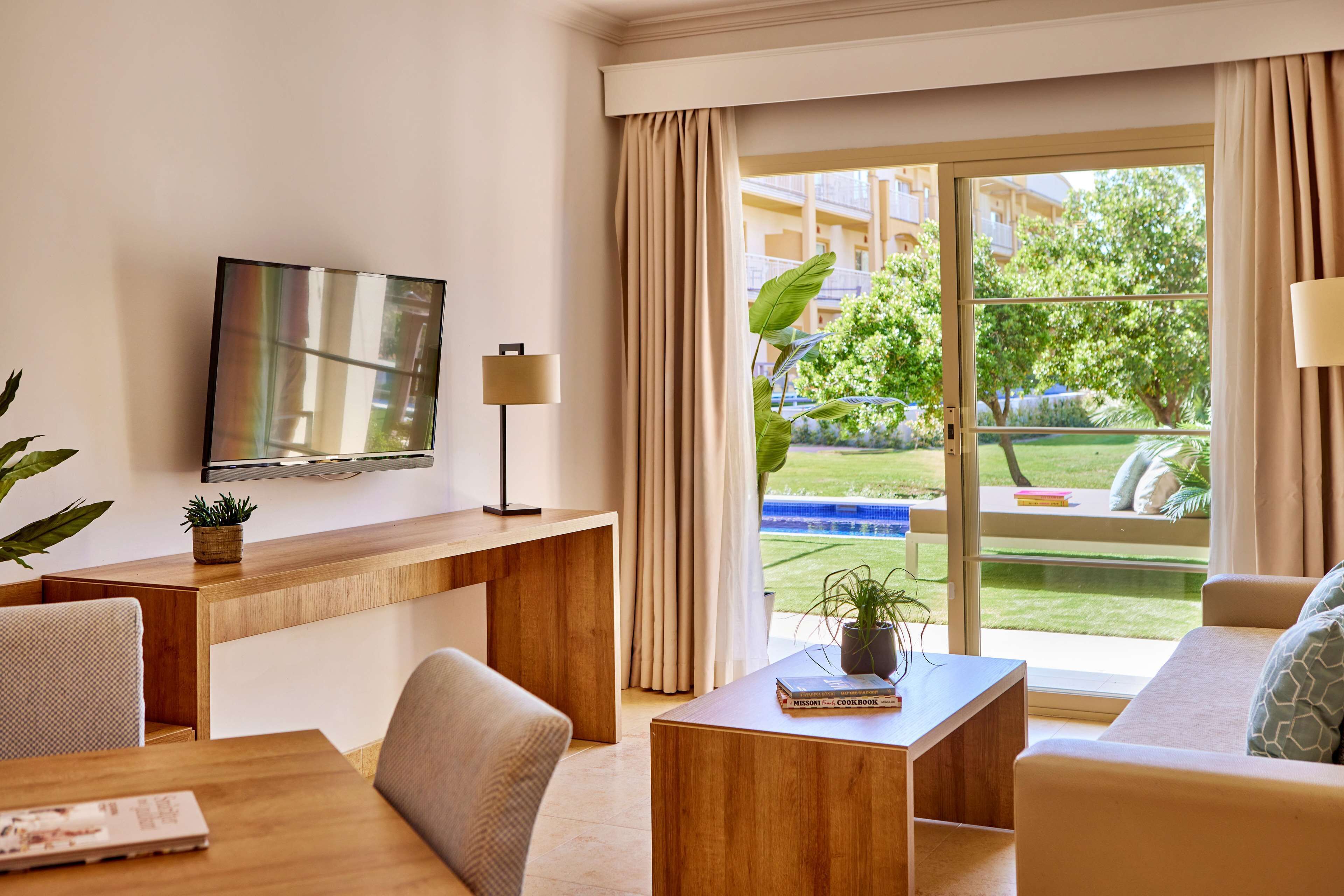 Ein modernes Wohnzimmer mit Schreibtisch aus Holz, einem Flachbildfernseher, einer Couch und Blick auf einen Garten durch eine Glastür.