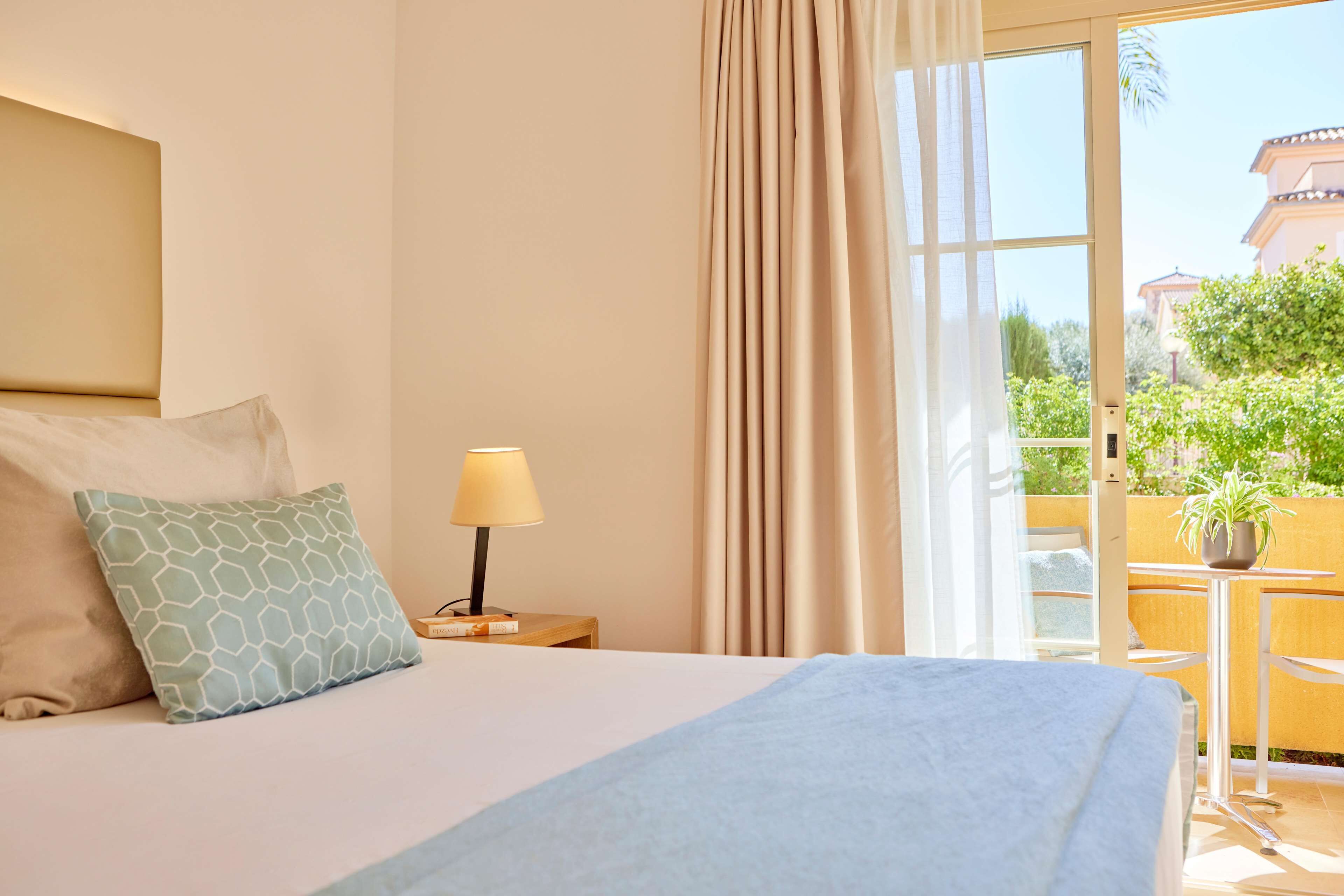 Ein Hotelzimmer mit Bett, Nachttisch mit Lampe, Fenster mit Vorhängen und Blick ins Freie.