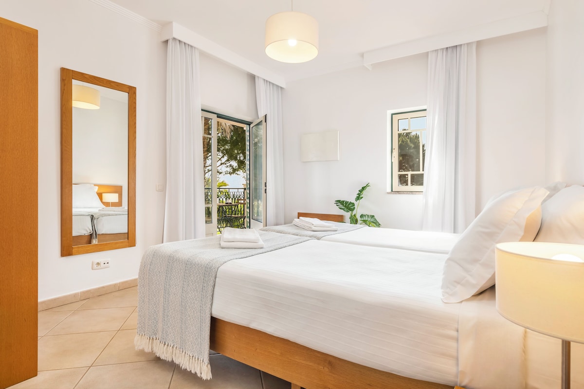 Habitación de hotel con una cama grande, una mesita de noche con lámpara, una ventana con cortinas y vistas al exterior.