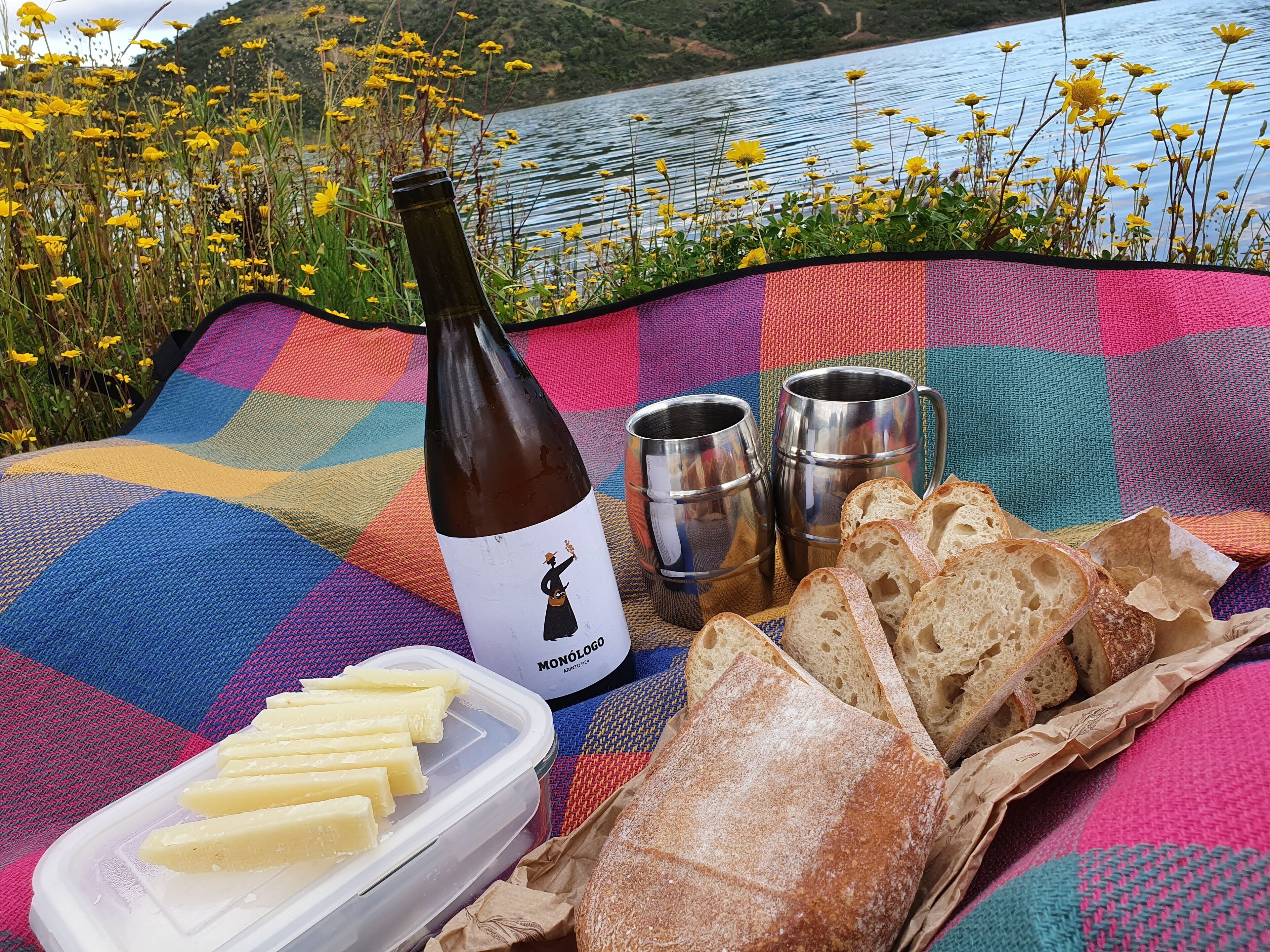Un pique-nique installé sur une couverture colorée au bord d’un lac, avec une bouteille de vin Monologo, deux mugs en acier inoxydable, un pot de beurre et une miche de pain.
