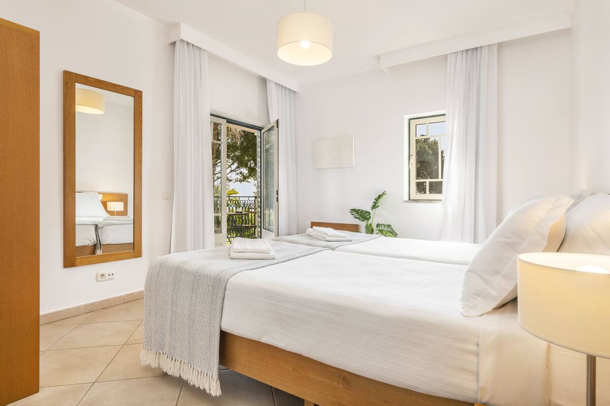 A hotel room with a large bed, a bedside table with a lamp, a window with curtains, and a view of the outside.