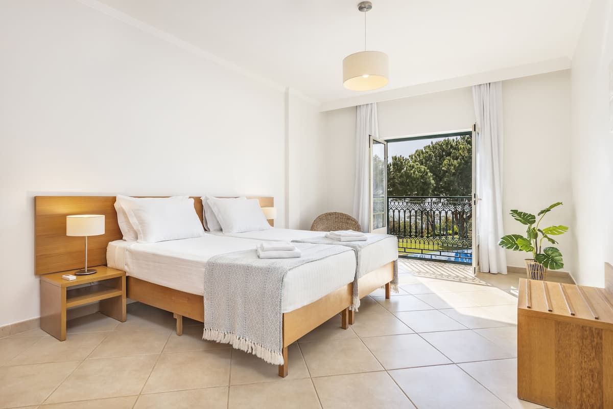 A modern hotel room with a double bed, a bedside table with a lamp, a small side table, a large window with a view of greenery, and a balcony with a railing.