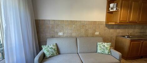 A neutral-toned sofa with two pillows, a kitchen area with wooden cabinets, and a window with white curtains.