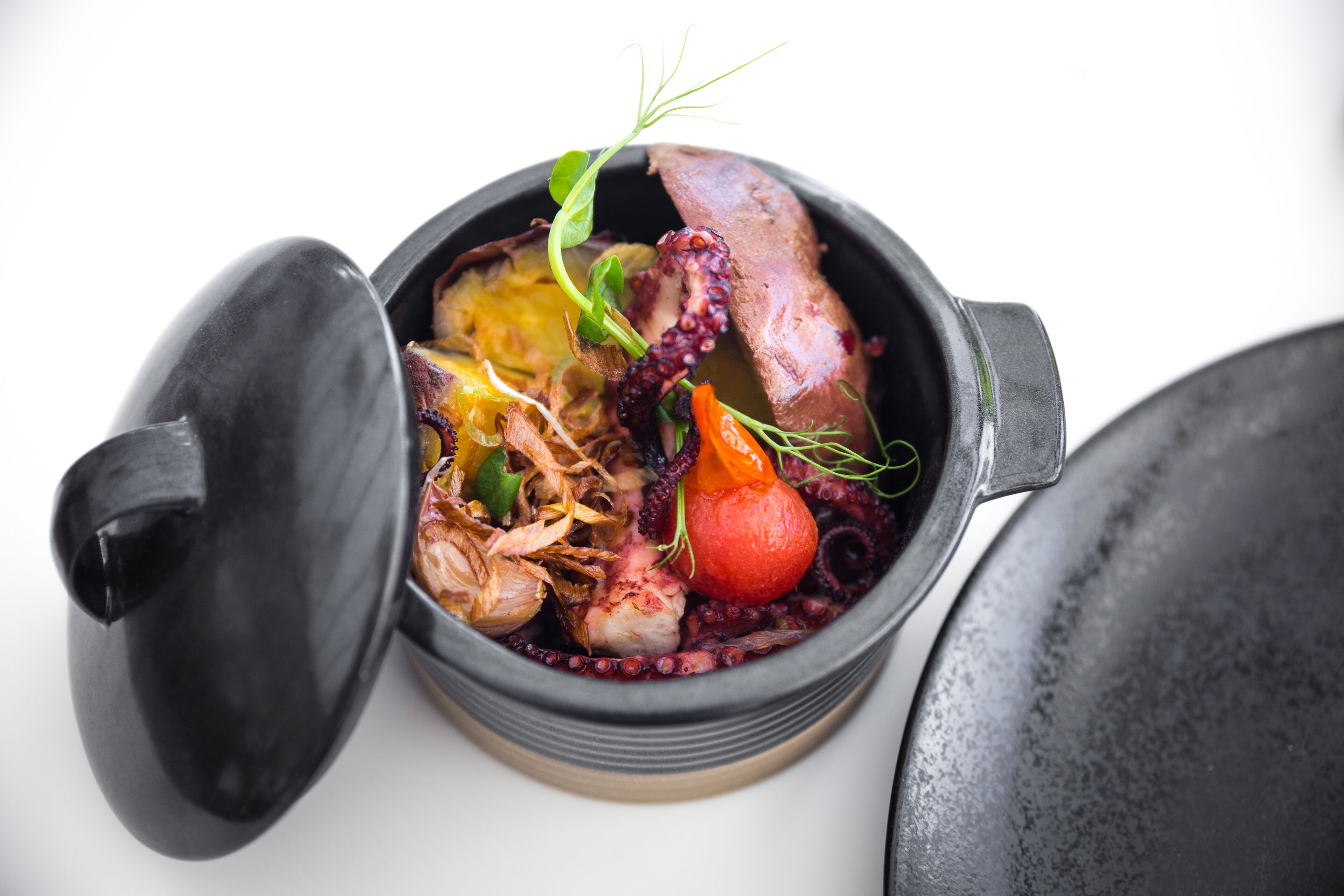 Un plat contenant des morceaux de poulpe, des légumes et un fruit rouge, servi dans un bol noir muni d’un couvercle.