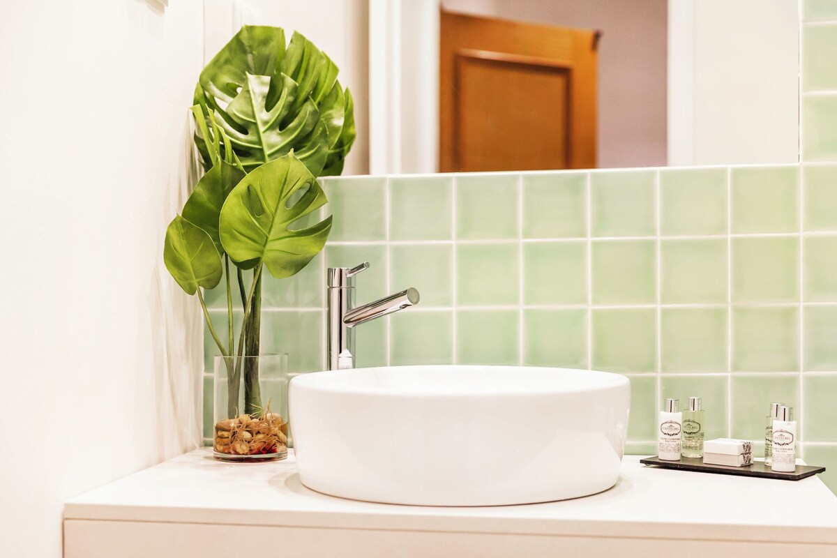 Un baño con un lavamanos blanco, azulejos verdes, una planta en maceta y artículos de aseo sobre el mostrador.