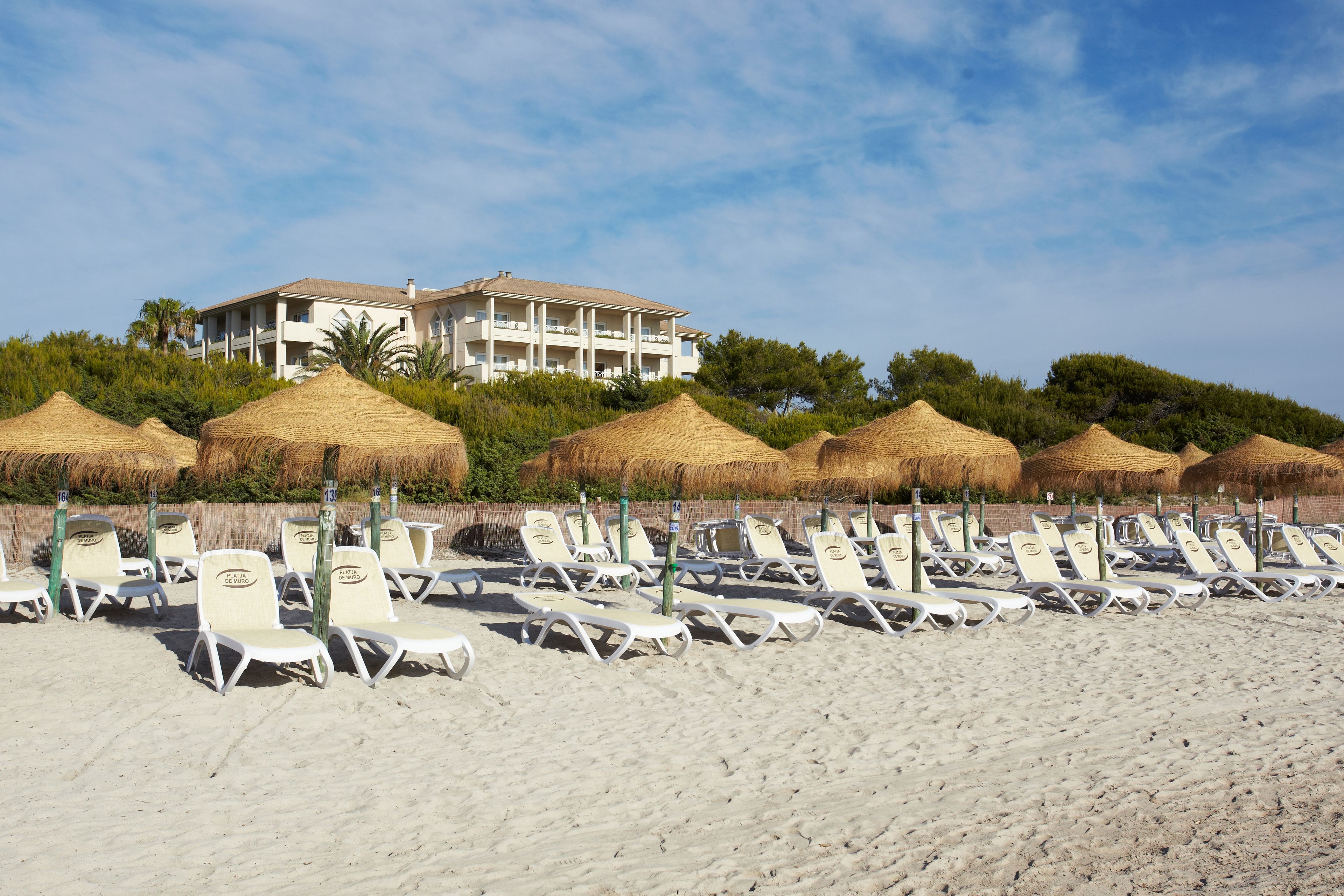 Ein Strand mit Reihen aus weißen Liegestühlen und strohgedeckten Sonnenschirmen, ein großes Gebäude im Hintergrund und ein klarer Himmel.