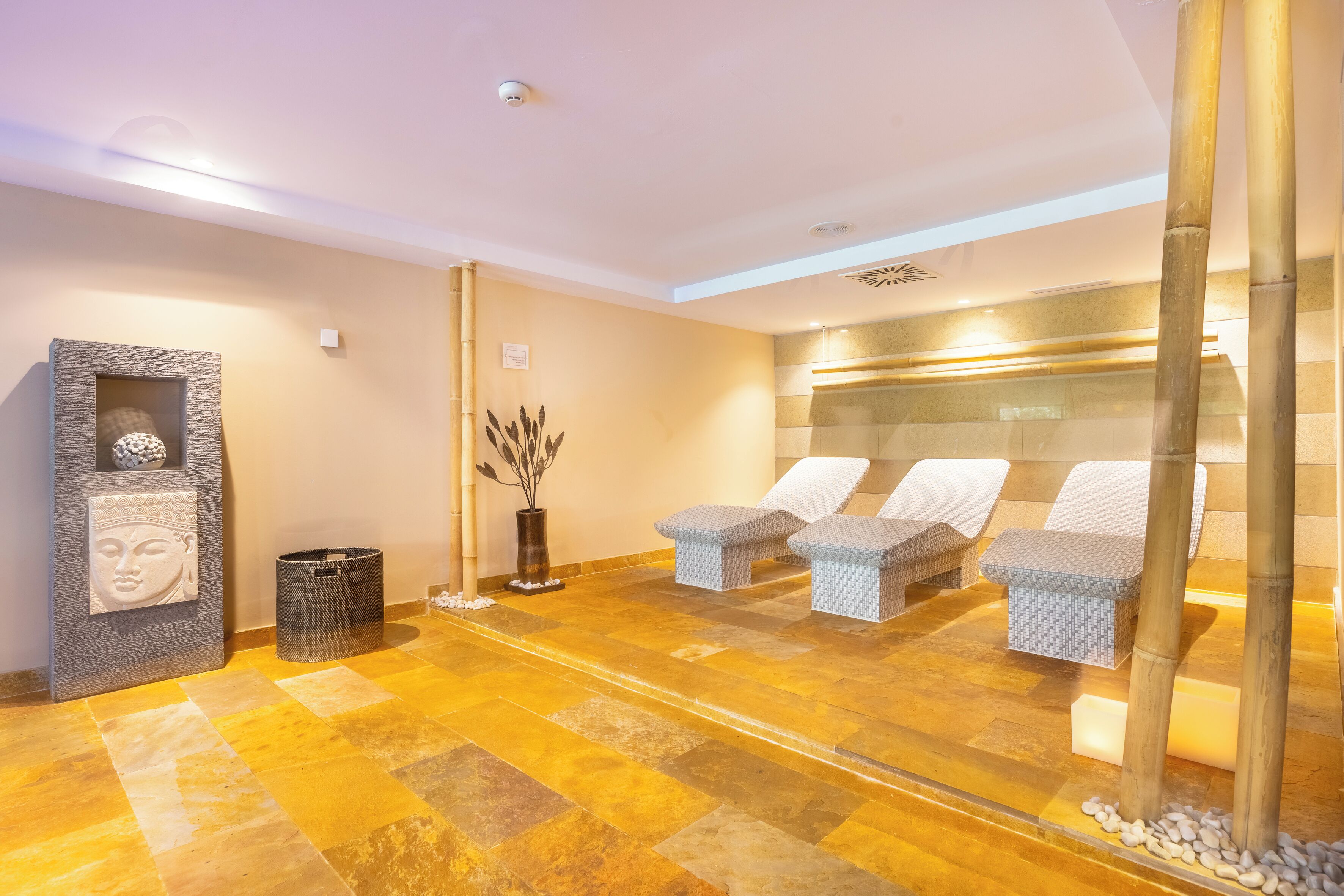 Zona de spa con suelo de baldosas, una camilla de masaje fijada a la pared, un cubo de basura y un jarrón decorativo con plantas secas.