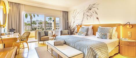 A hotel room with a large bed, a desk, a chair, a bench, and a view of palm trees and buildings.