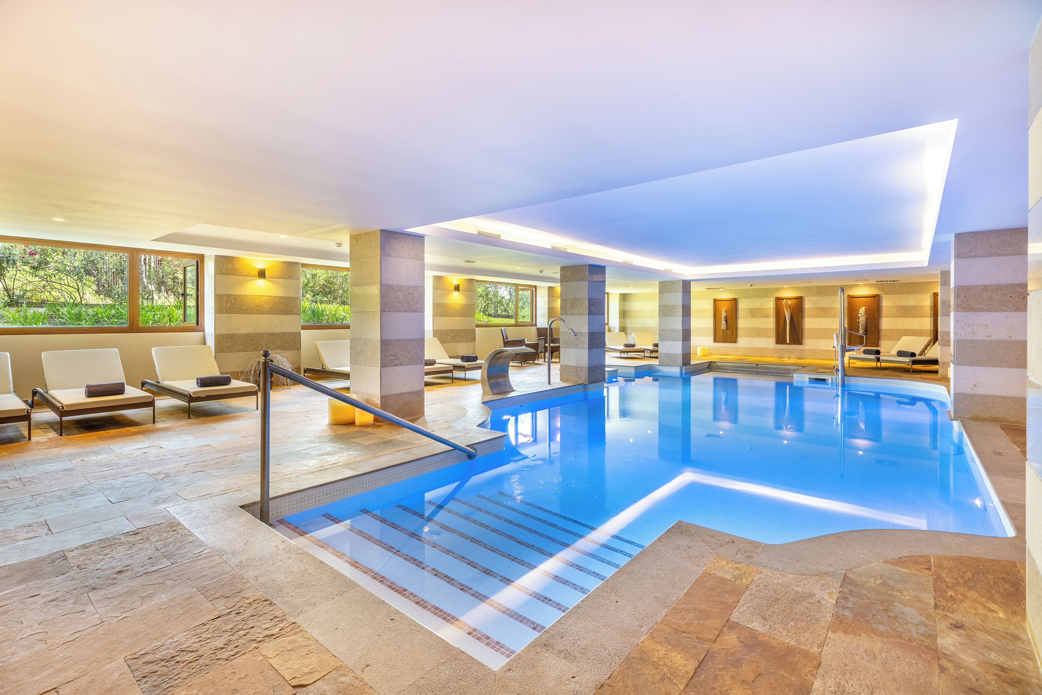 A modern indoor swimming pool with clear water, lounge chairs, and a view of greenery outside.