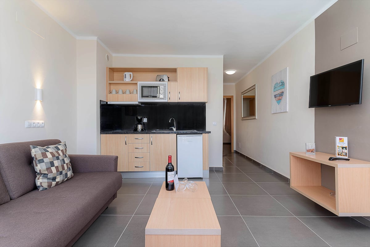 A modern living room with a sofa, a coffee table, a TV, and a kitchen area with cabinets and appliances.