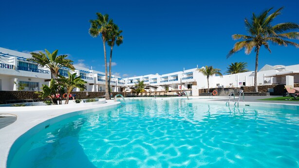 A large swimming pool with clear blue water, surrounded by white buildings with blue accents and palm trees. - Hotel Siroco - Adults Only (Teguise)