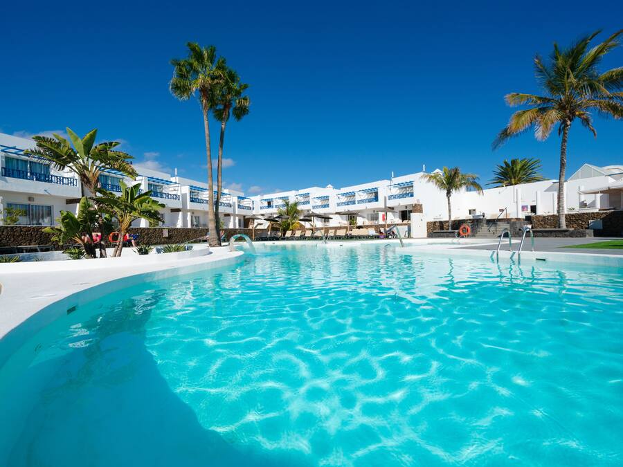 A large swimming pool with clear blue water, surrounded by white buildings with blue accents and palm trees.