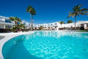 A large swimming pool with clear blue water, surrounded by white buildings with blue accents and palm trees. - Hotel Siroco - Adults Only (Teguise)