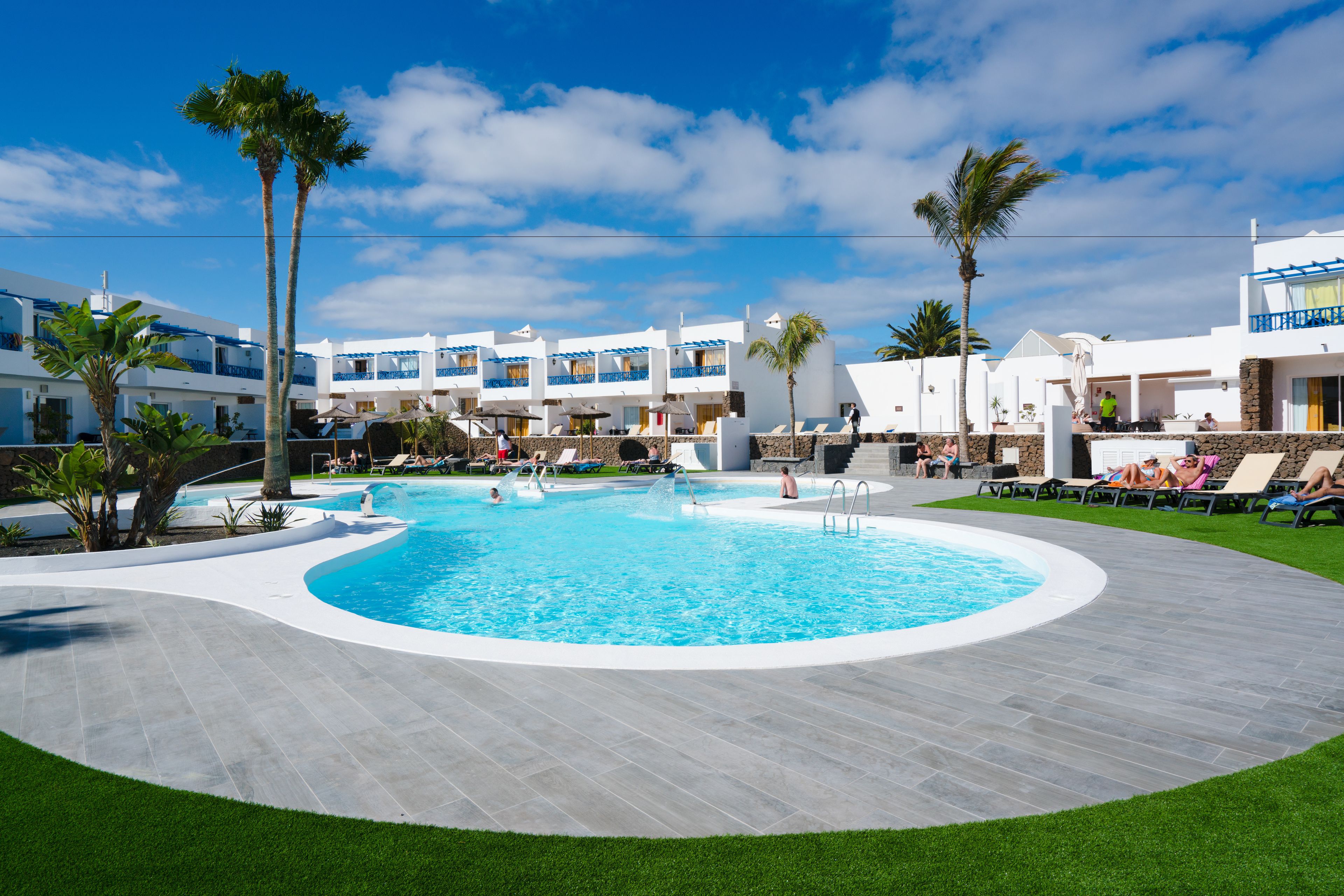 Una piscina rodeada por un edificio blanco con balcones, palmeras y un cielo azul despejado.