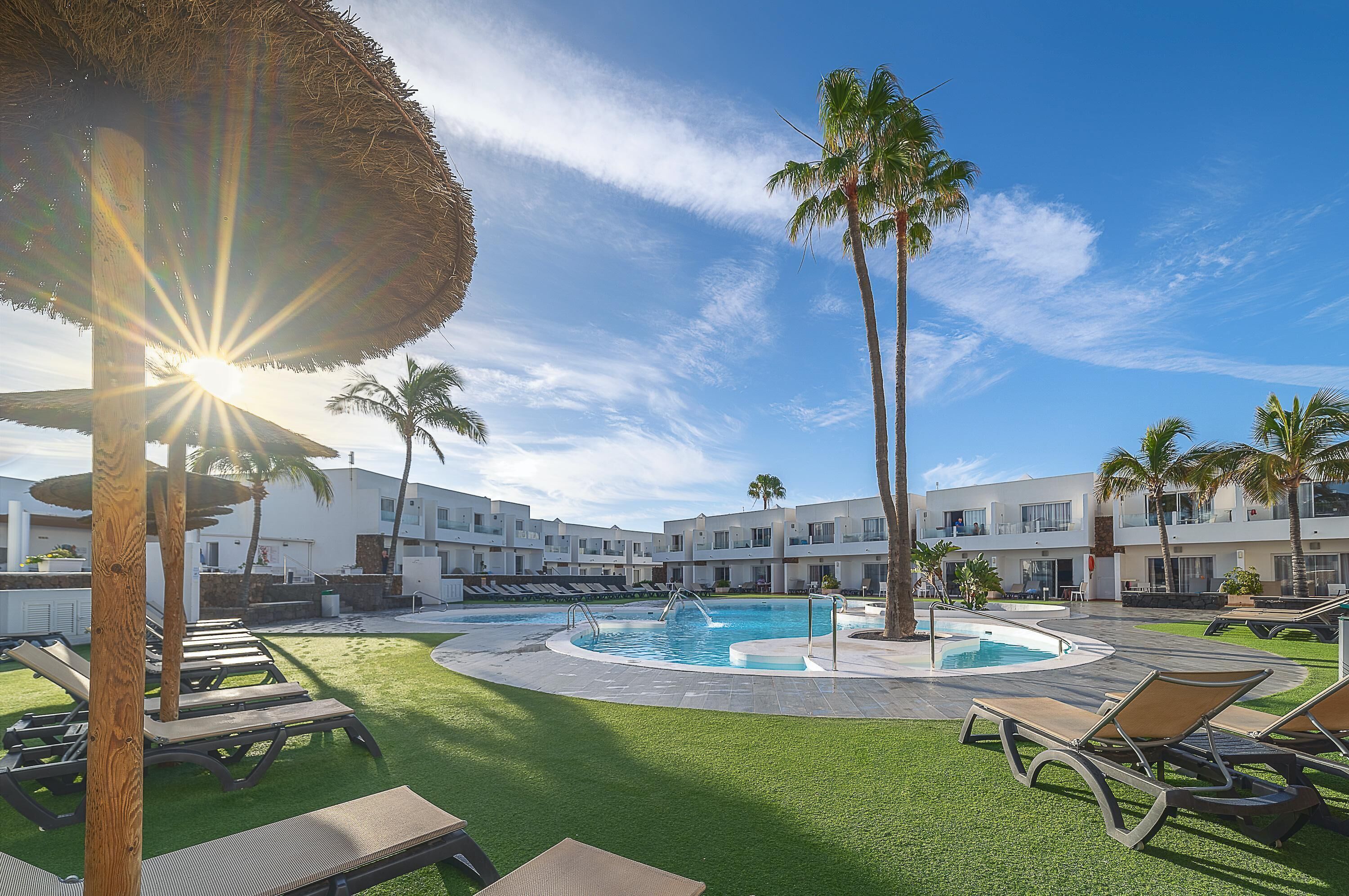 Zona de piscina con sillas de descanso, palmeras y un edificio con balcones.
