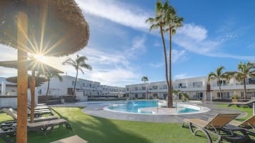 Zona de piscina con sillas de descanso, palmeras y un edificio con balcones.